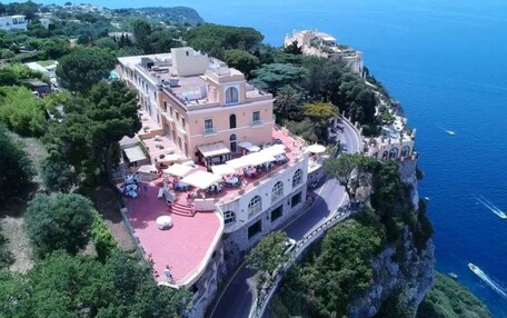 An aerial view of a coastal hotel with a red-roofed terrace, a stone facade, and a view of the sea.
