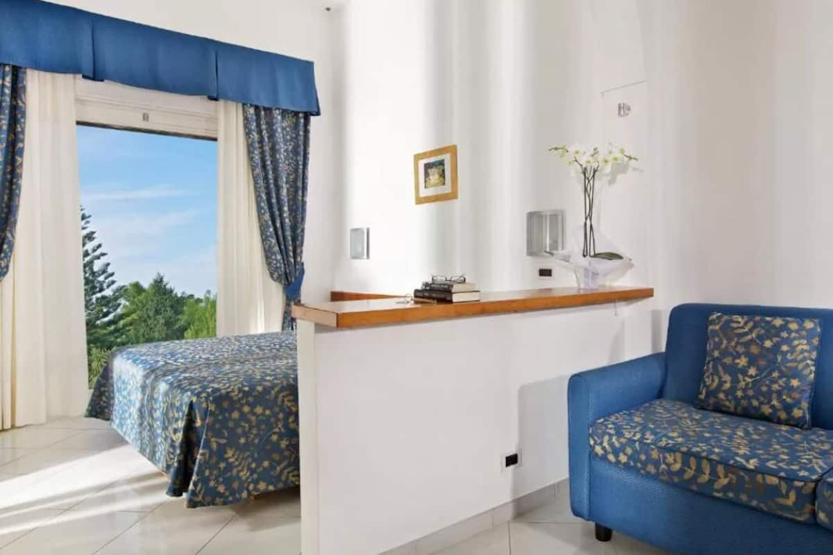 Ein Hotelzimmer mit zwei Betten, einem Schlafsofa, einem Holztisch, einer kleinen Ablage mit Büchern und einer Vase sowie einem Fenster mit Vorhängen.