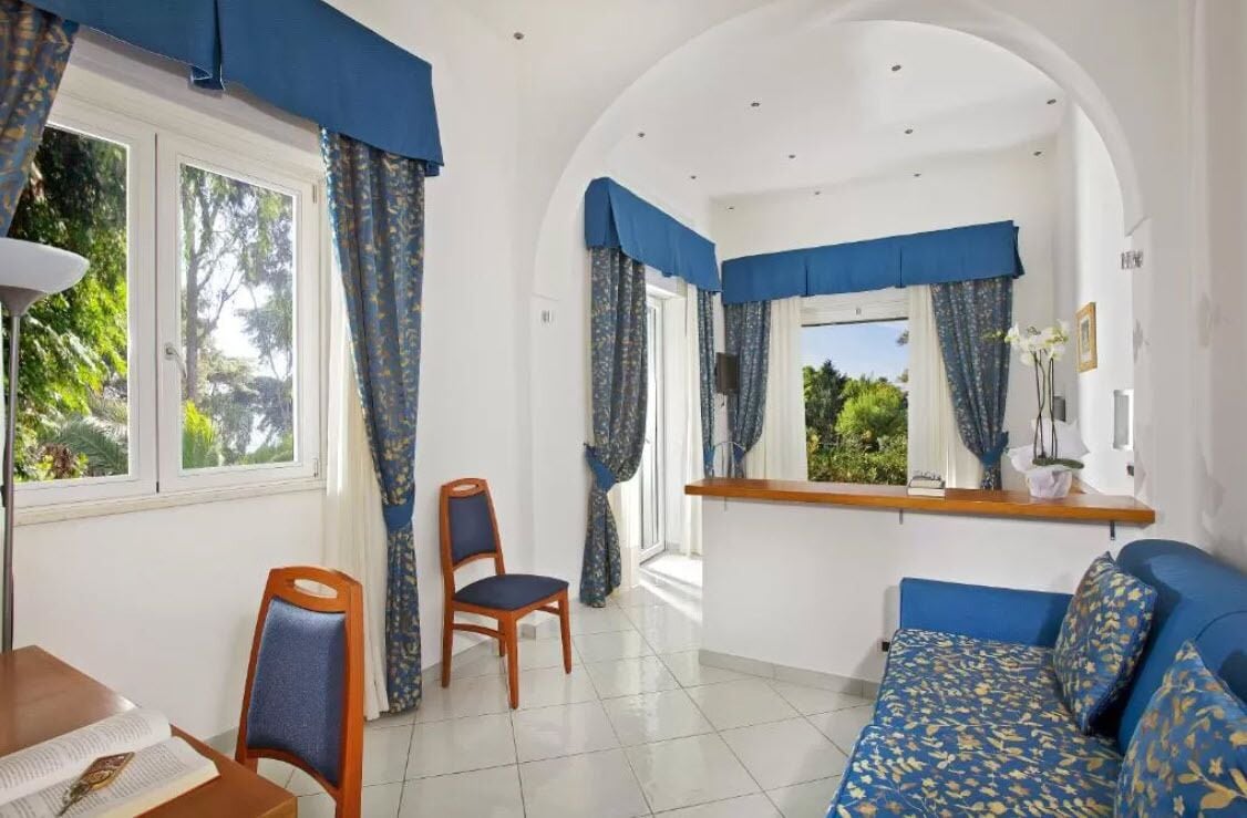 A room with a sofa, two chairs, a table with a book, and a window with curtains.