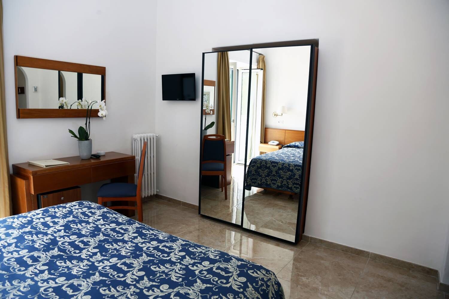 A bedroom with a bed, a desk, a chair, a mirror, and a television.