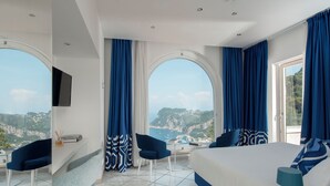 A modern hotel room with a large bed, two chairs, a desk, and a TV. The room has large windows with blue curtains and a view of a coastal landscape. - Hotel San Michele (Anacapri)