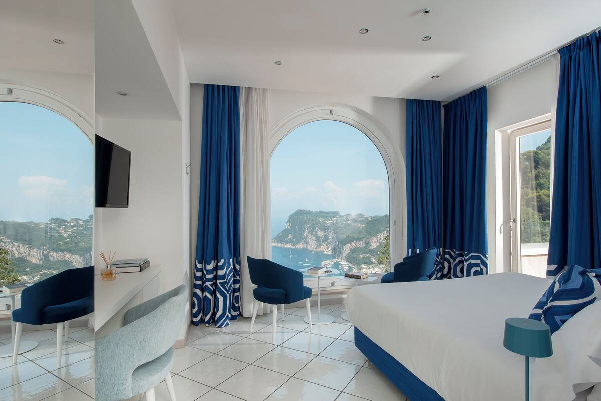Ein modernes Hotelzimmer mit einem großen Bett, zwei Sesseln, einem Schreibtisch und einem Fernseher. Das Zimmer verfügt über große Fenster mit blauen Vorhängen und Blick auf eine Küstenlandschaft.