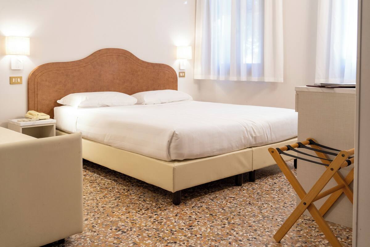 A hotel room with a bed, bedside lamps, a small table, a chair, and a window with curtains.