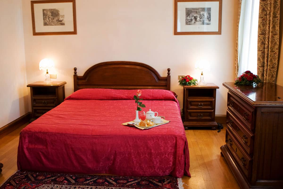 A neatly made bed with a red bedspread and a tray of breakfast items. There are two bedside tables with lamps, a dresser, and a patterned rug on the floor.