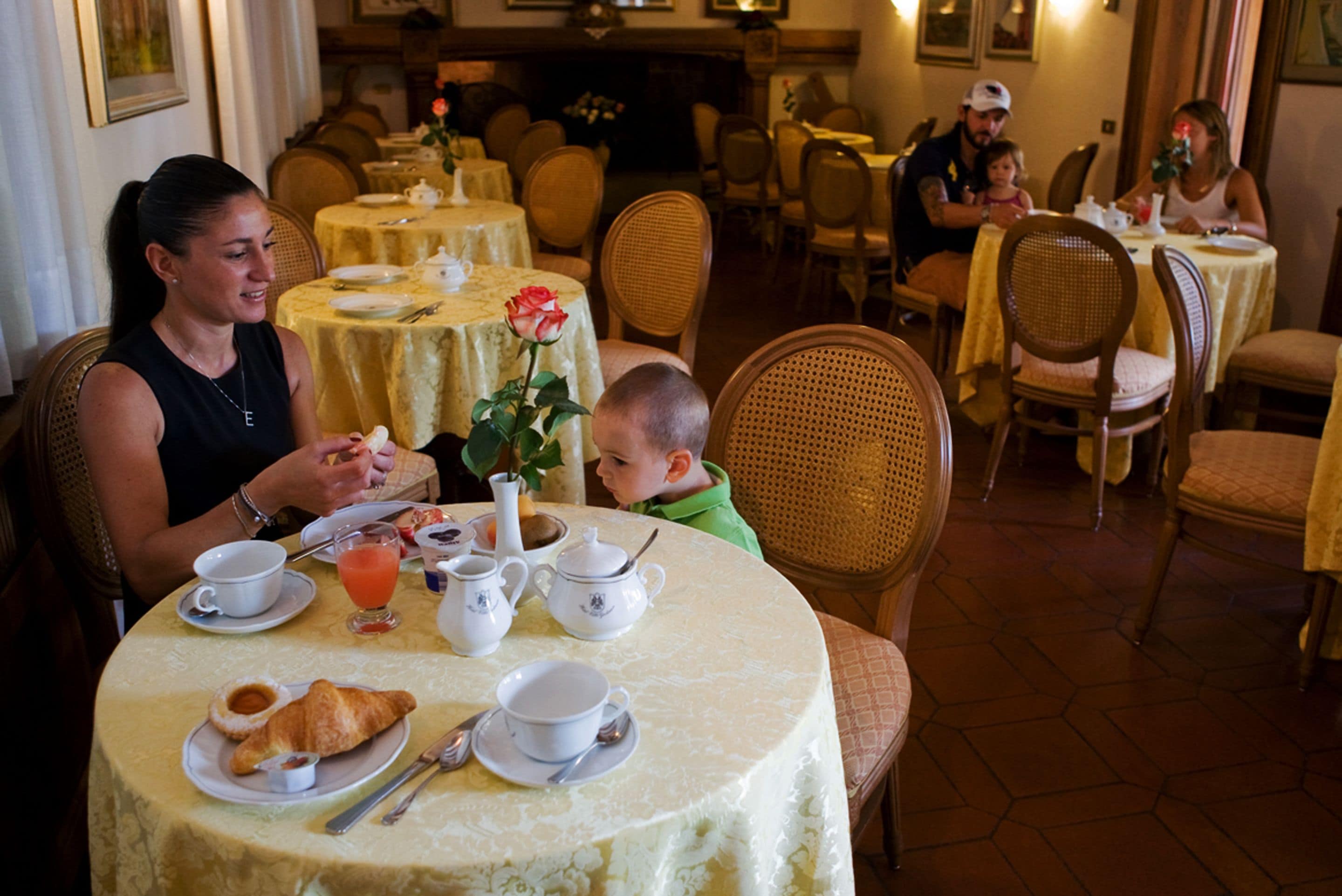Un comedor con mesas redondas cubiertas con manteles amarillos, sillas y una variedad de vajilla, incluyendo tazas, platos y un centro de mesa con rosas.