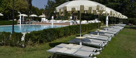 Zona junto a la piscina con sillones reclinables y sombrillas, rodeada de vegetación.