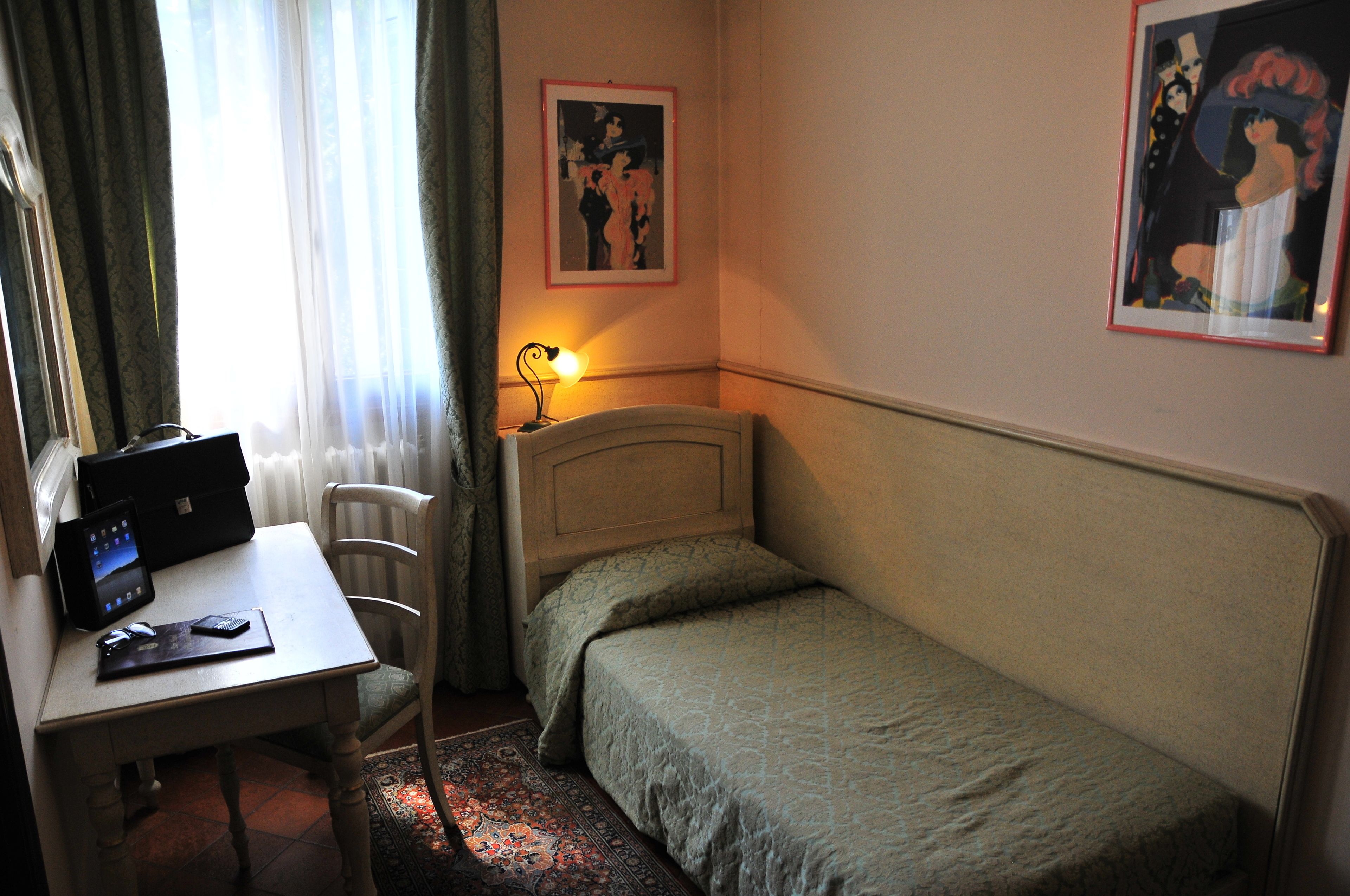 A bedroom with a bed, a desk with a computer and mouse, a lamp, and two framed pictures on the wall.