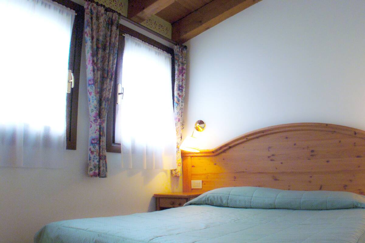 A bedroom with a wooden headboard, a single bed, a nightstand with a lamp, and a window with curtains.