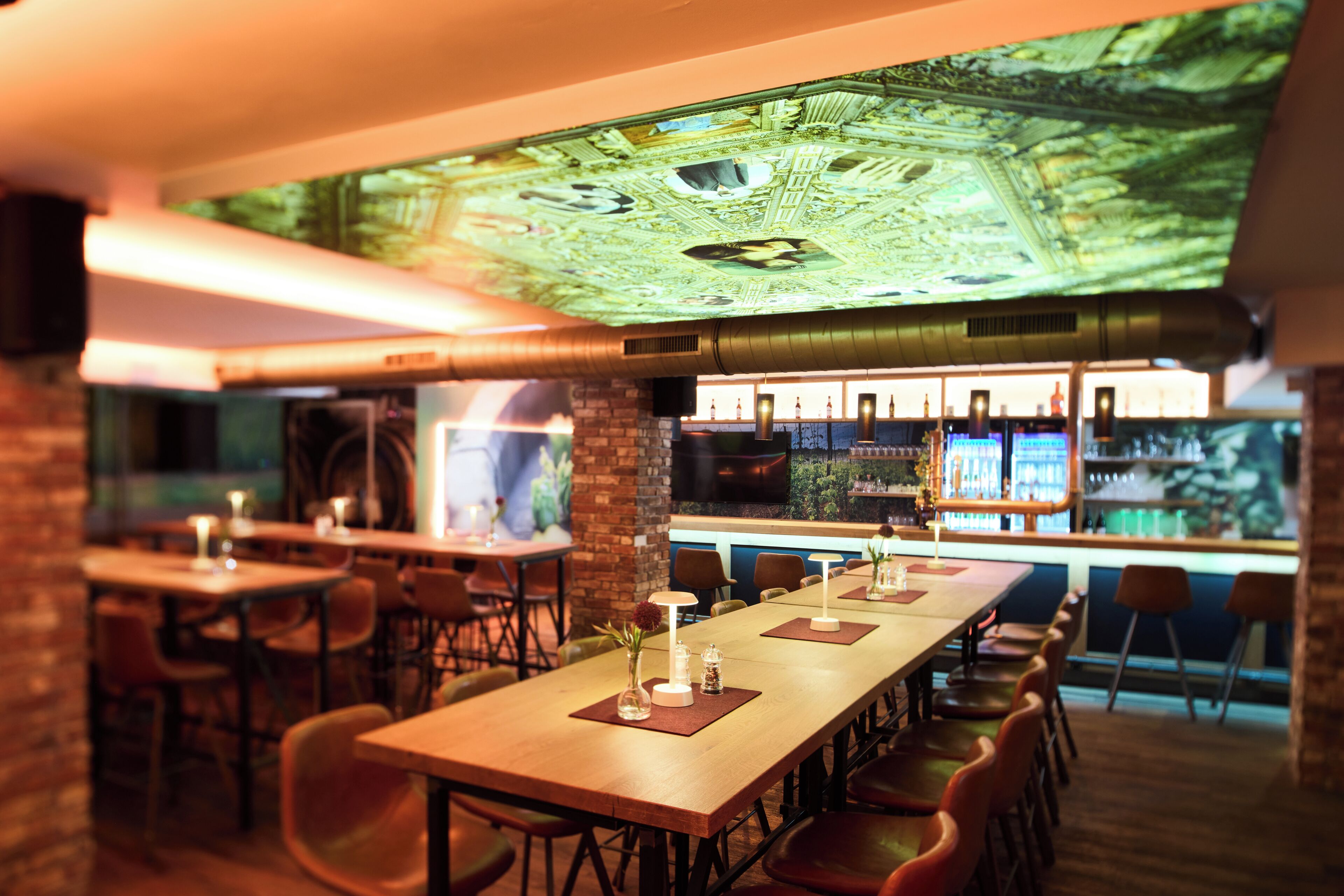 A bar area with long wooden tables, bar stools, and a large screen displaying an image.