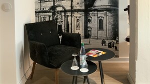 Una habitación con un mural de una iglesia, una silla negra, una mesita redonda con una botella y vasos, y un libro.