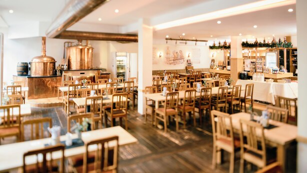 Un interior de restaurante con mesas y sillas de madera, una zona de bar y un gran equipo de elaboración de cerveza de cobre.