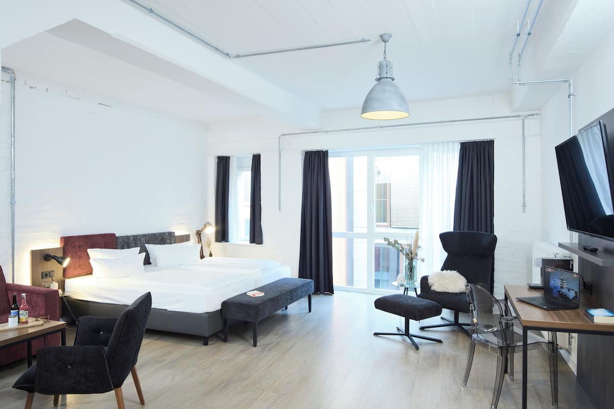 A modern hotel room with a large bed, a desk with a laptop, a TV, and a seating area with chairs and a bench.