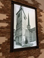 Fotografia em preto e branco emoldurada de um edifício histórico com uma torre e um pináculo imponentes, fixada numa parede de tijolos.