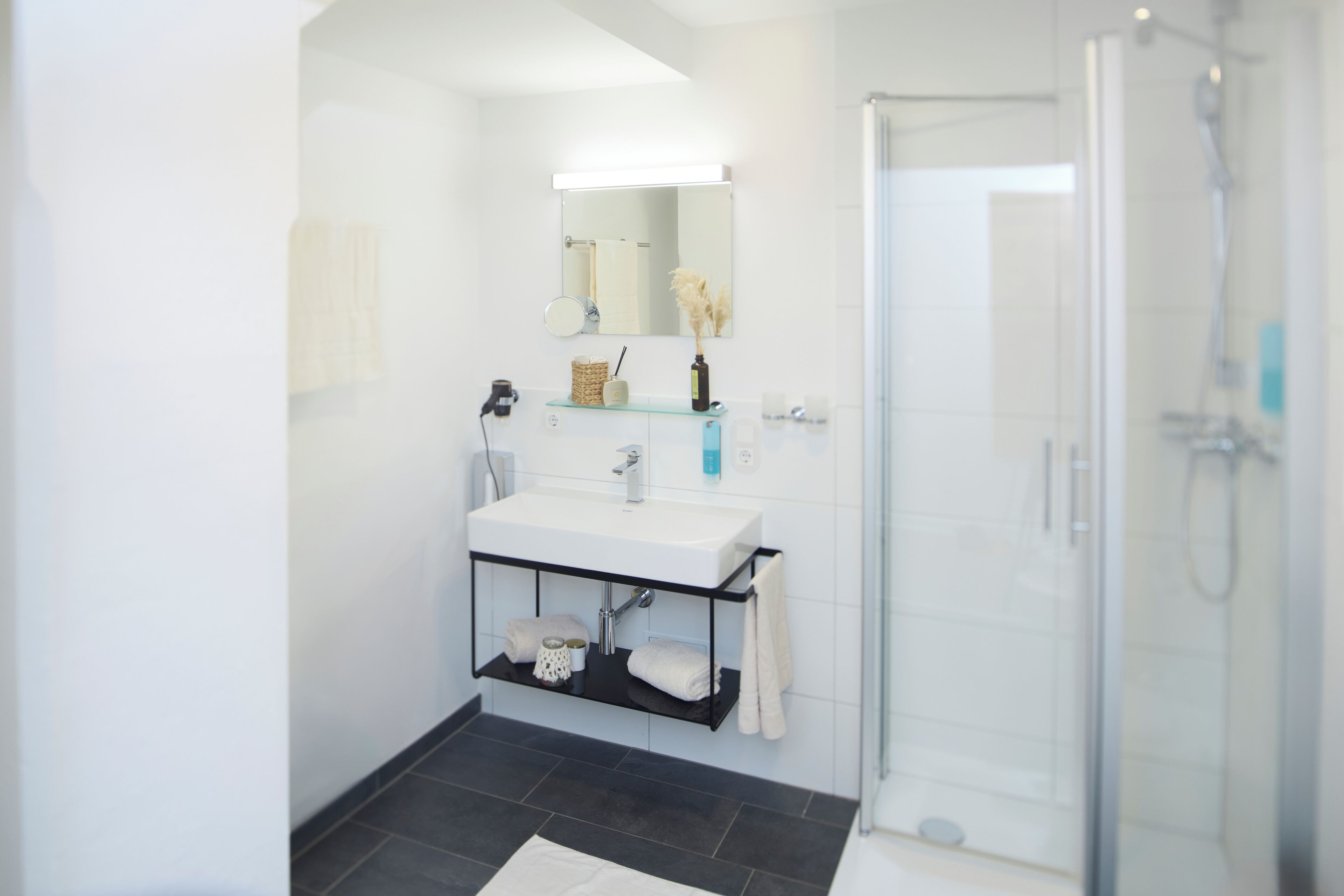 A modern bathroom with a white sink, black shelving, a glass shower enclosure, and a mirror above the sink.