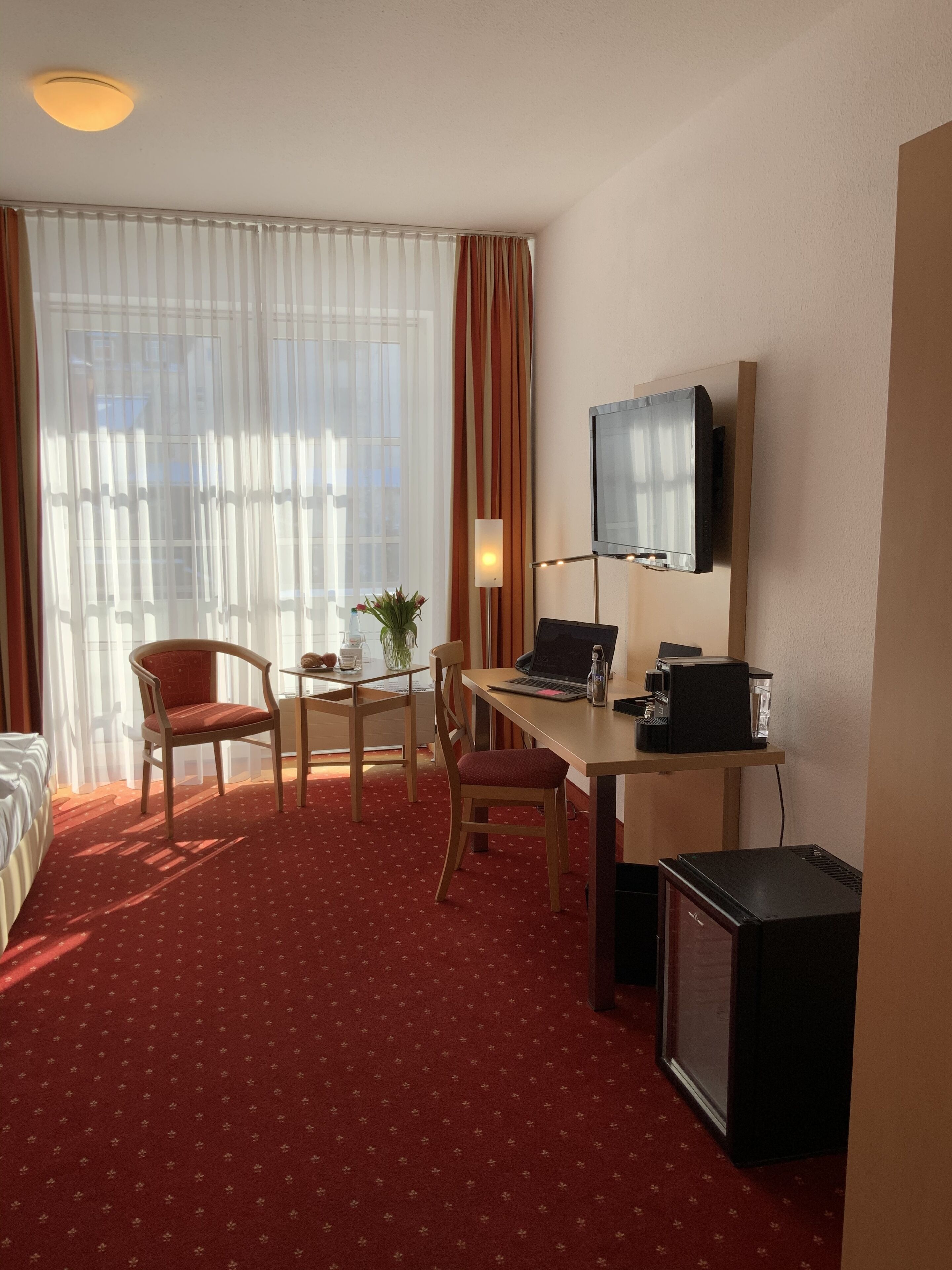 A hotel room with a bed, a desk with a laptop, a television, and a small refrigerator.