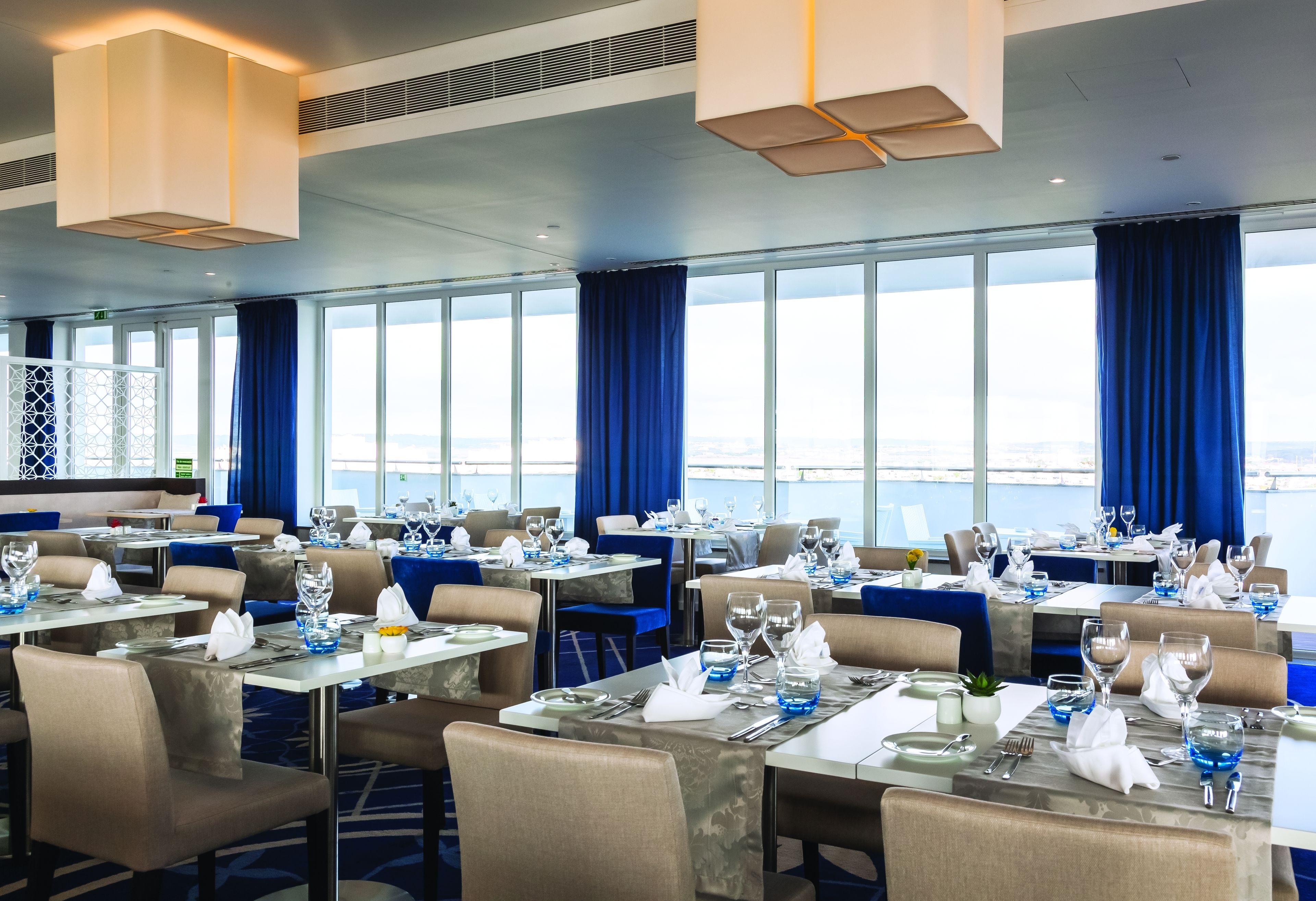 A restaurant with tables set for dining, large windows with curtains, and a view of the sea.