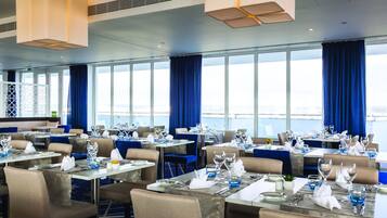 A restaurant with tables set for dining, large windows with curtains, and a view of the sea.