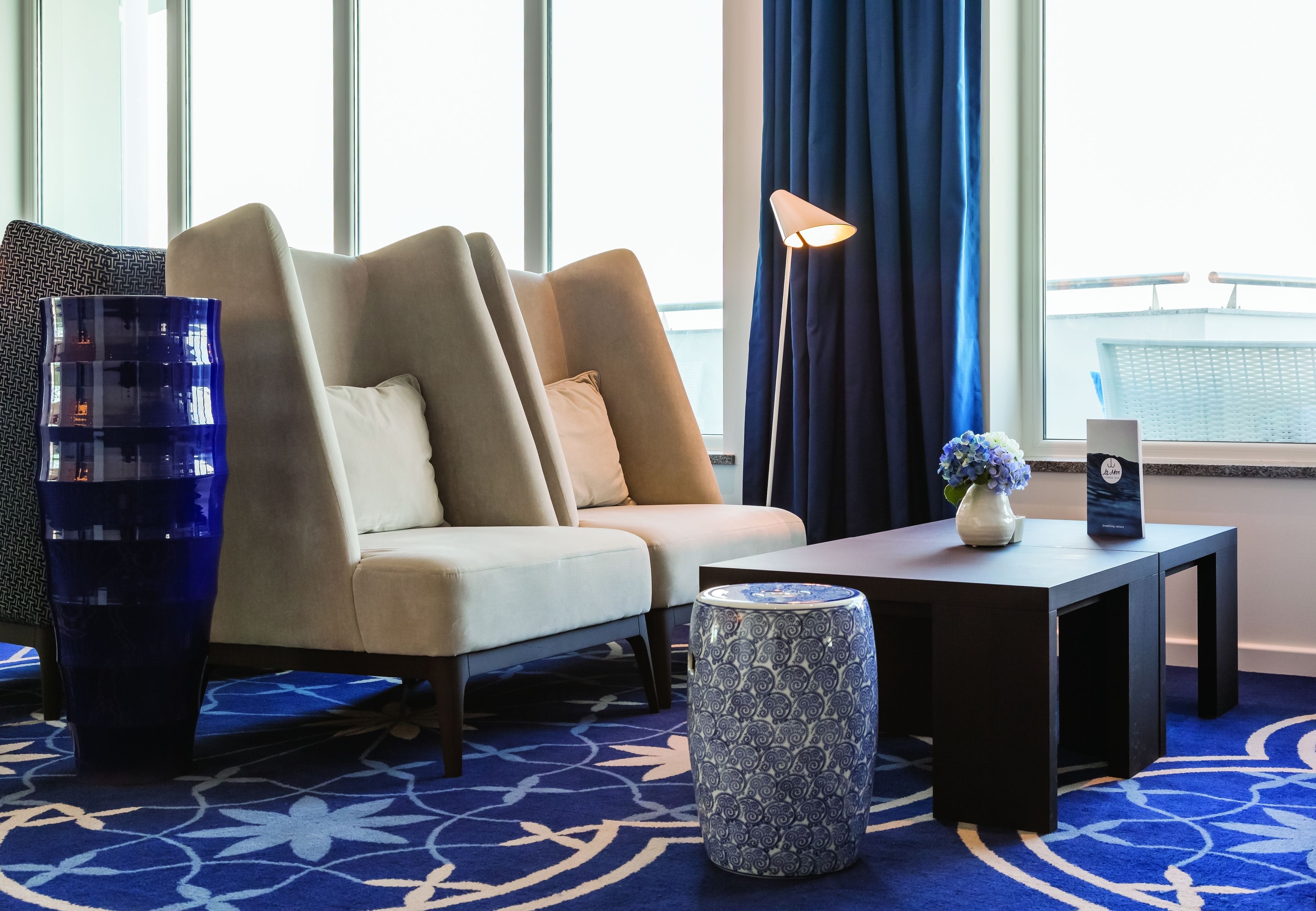 Een moderne hotelkamer met een beige bank, een donkerhouten salontafel, een decoratieve vaas en een kamerbreed tapijt met dessin.