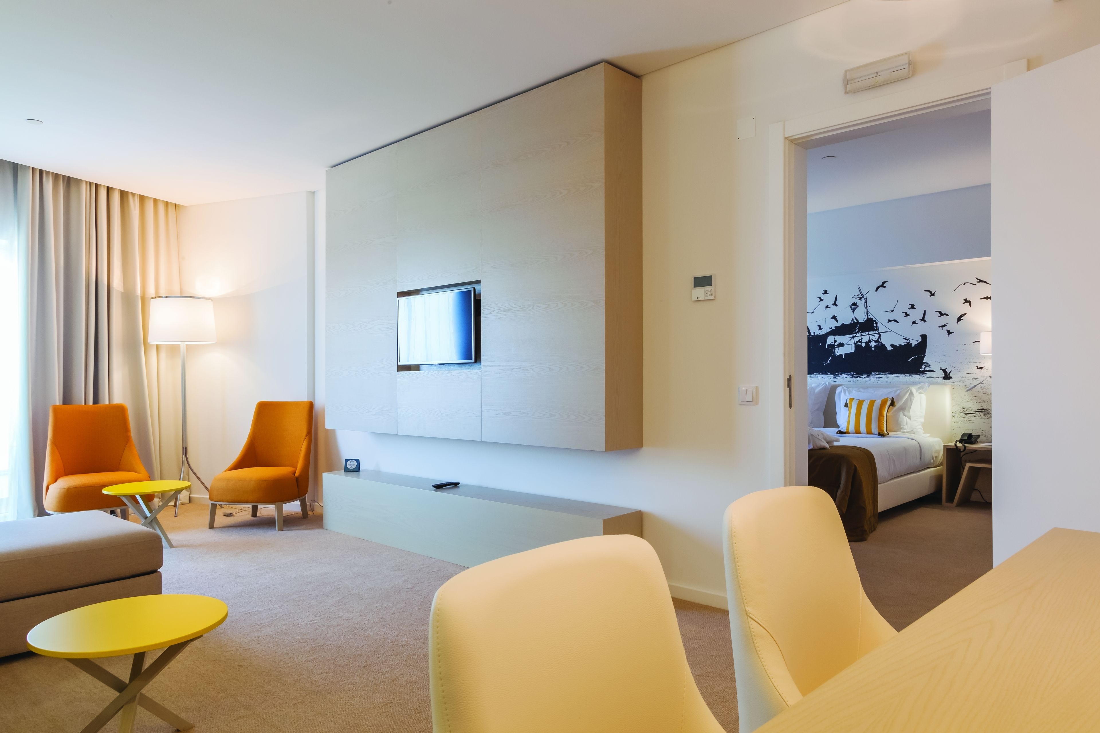 Een moderne hotelkamer met een sofa, twee fauteuils, een klein tafeltje, een bed met nachtkastje en een tv aan de muur.