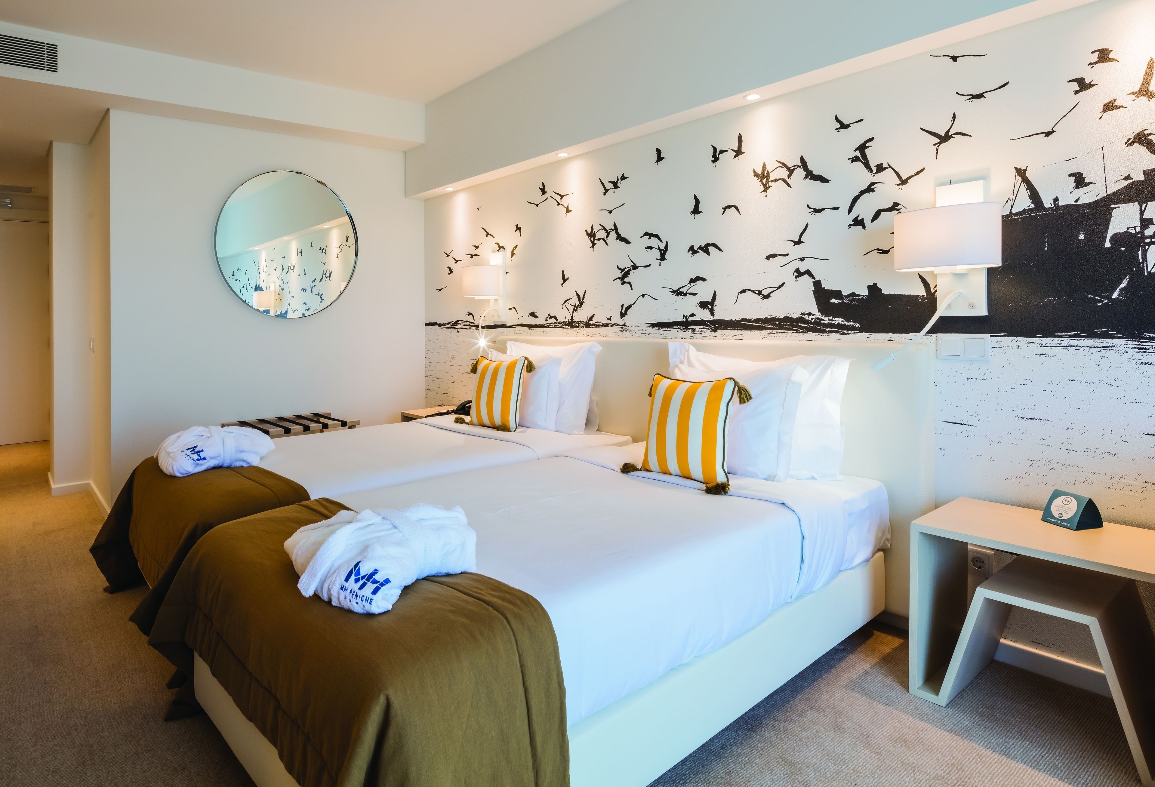 A hotel room with two beds, a round mirror, a wall mural of birds, and a bedside table with a lamp.