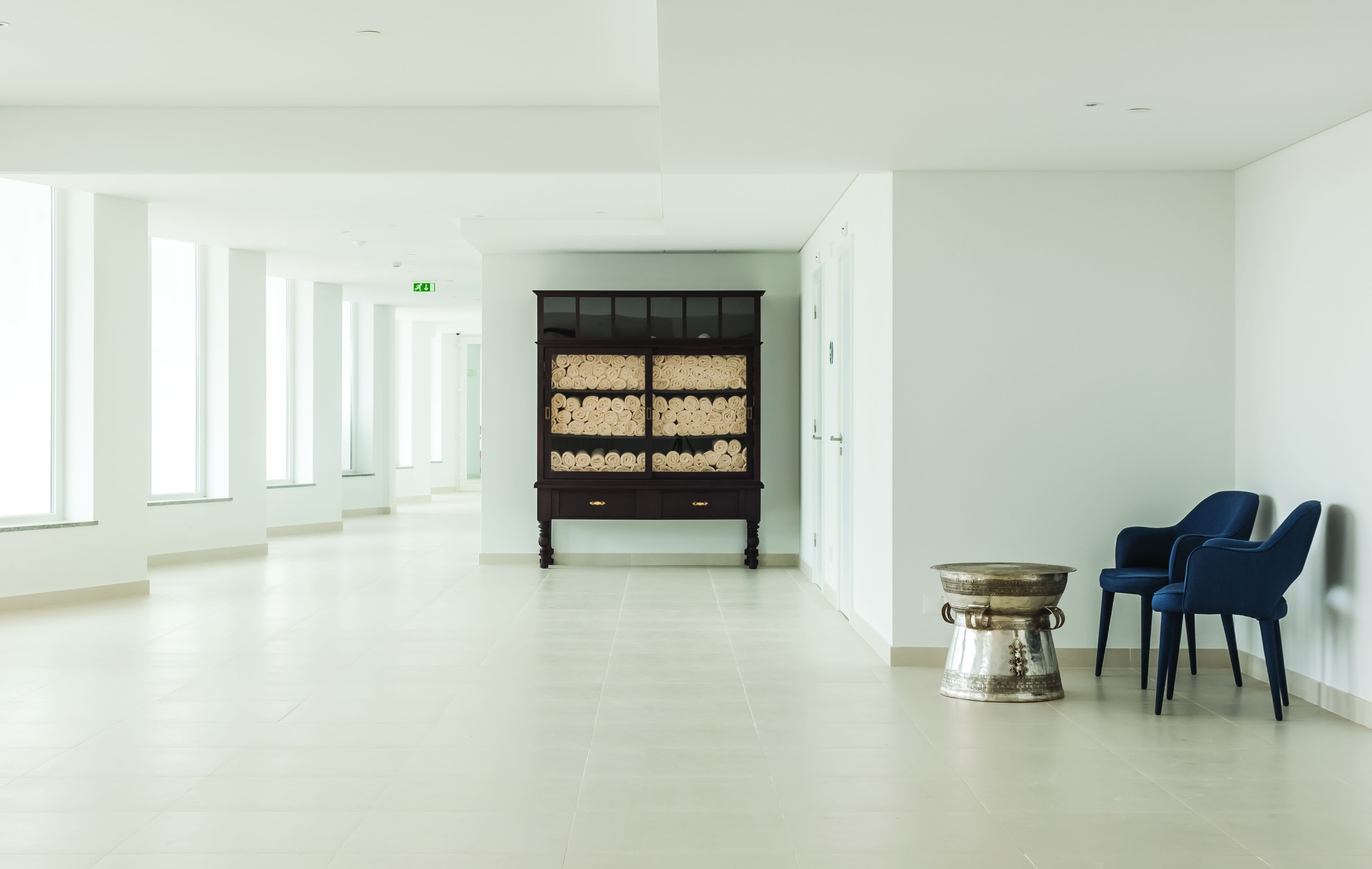A spacious, well-lit hallway with a large wooden cabinet, two blue chairs, and a metal table.