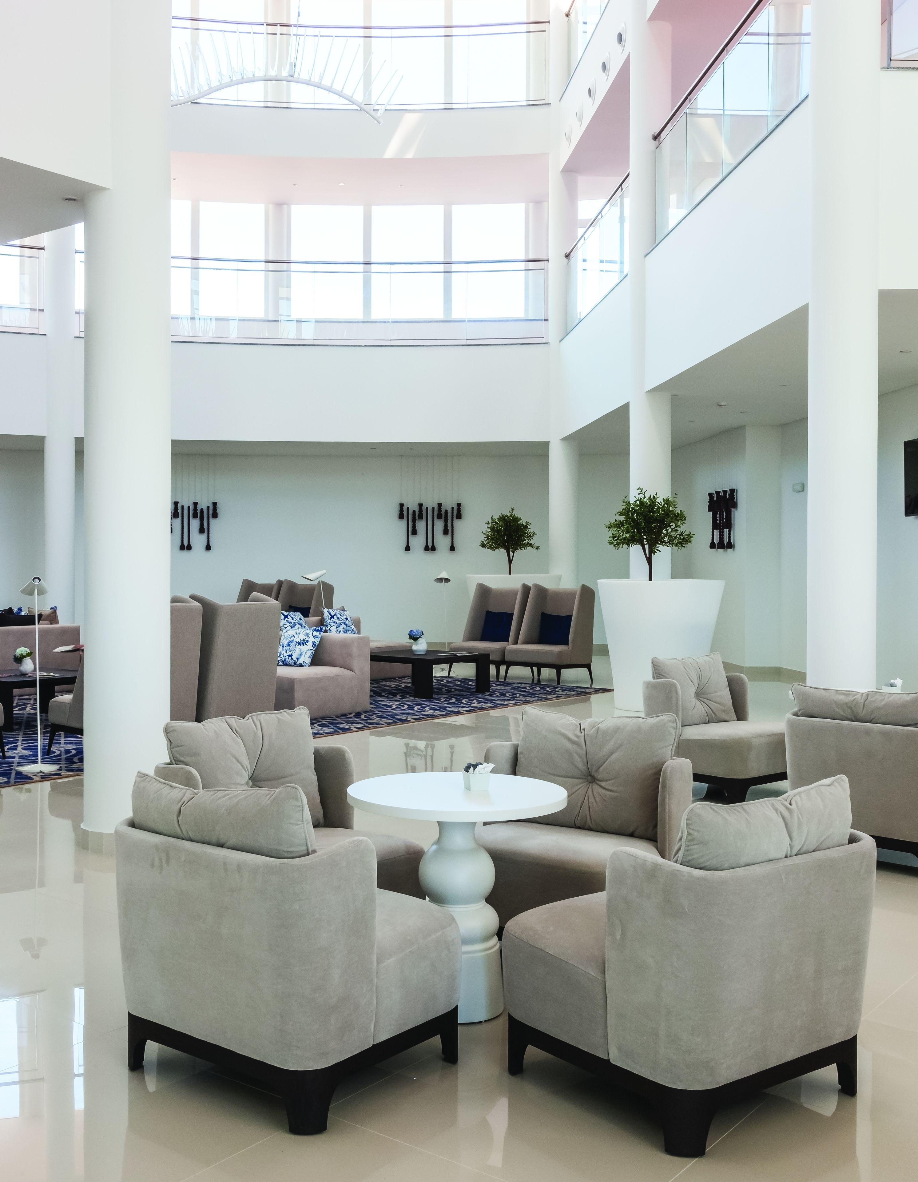 A spacious, modern lobby with high ceilings, a central seating area with sofas and armchairs, a round table, and a large window providing natural light.