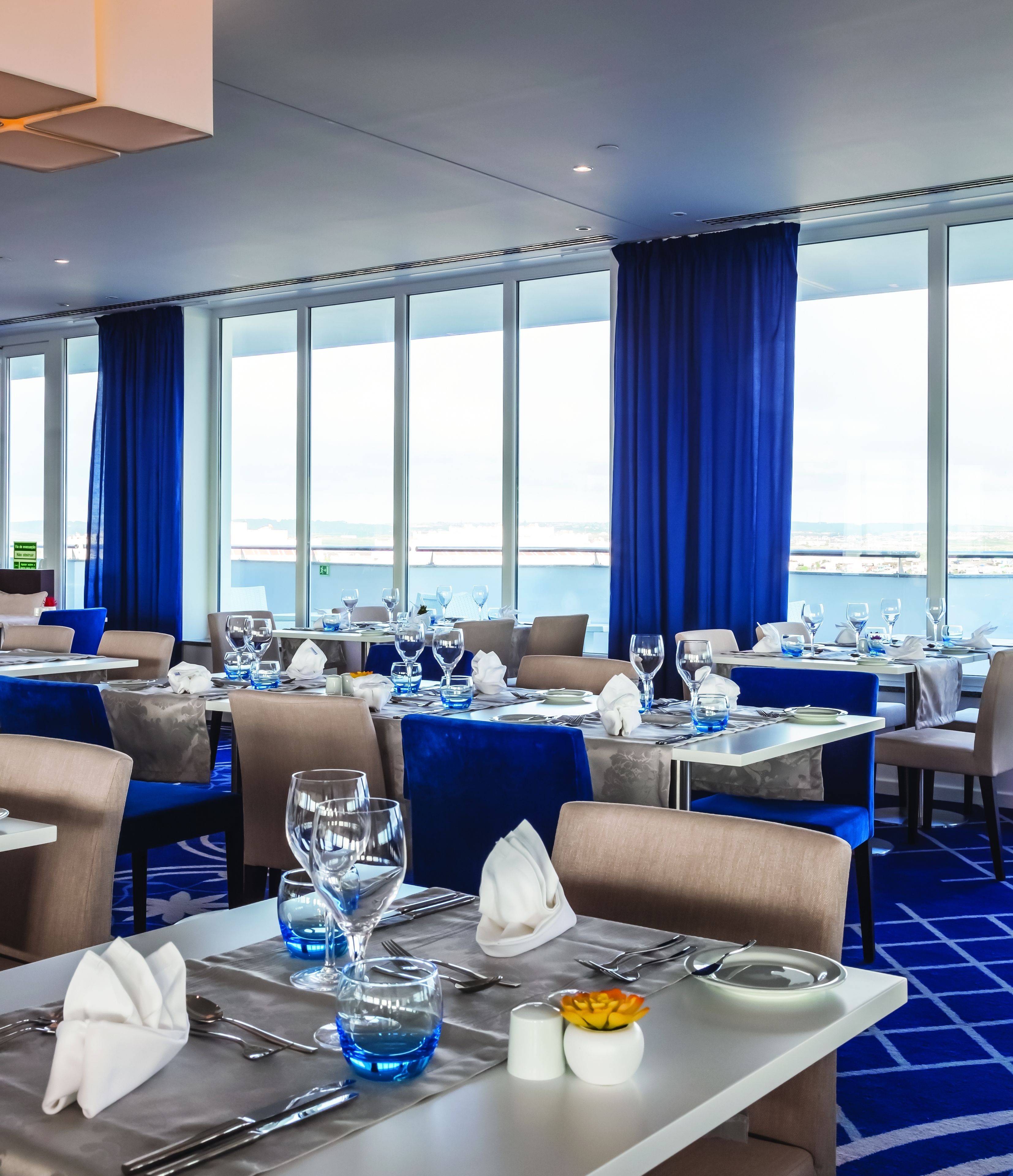 Comedor con mesas dispuestas para el servicio, ventanas amplias con cortinas azules y vista a la ciudad.