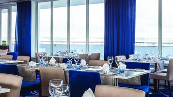 Comedor con mesas dispuestas para el servicio, ventanas amplias con cortinas azules y vista a la ciudad.