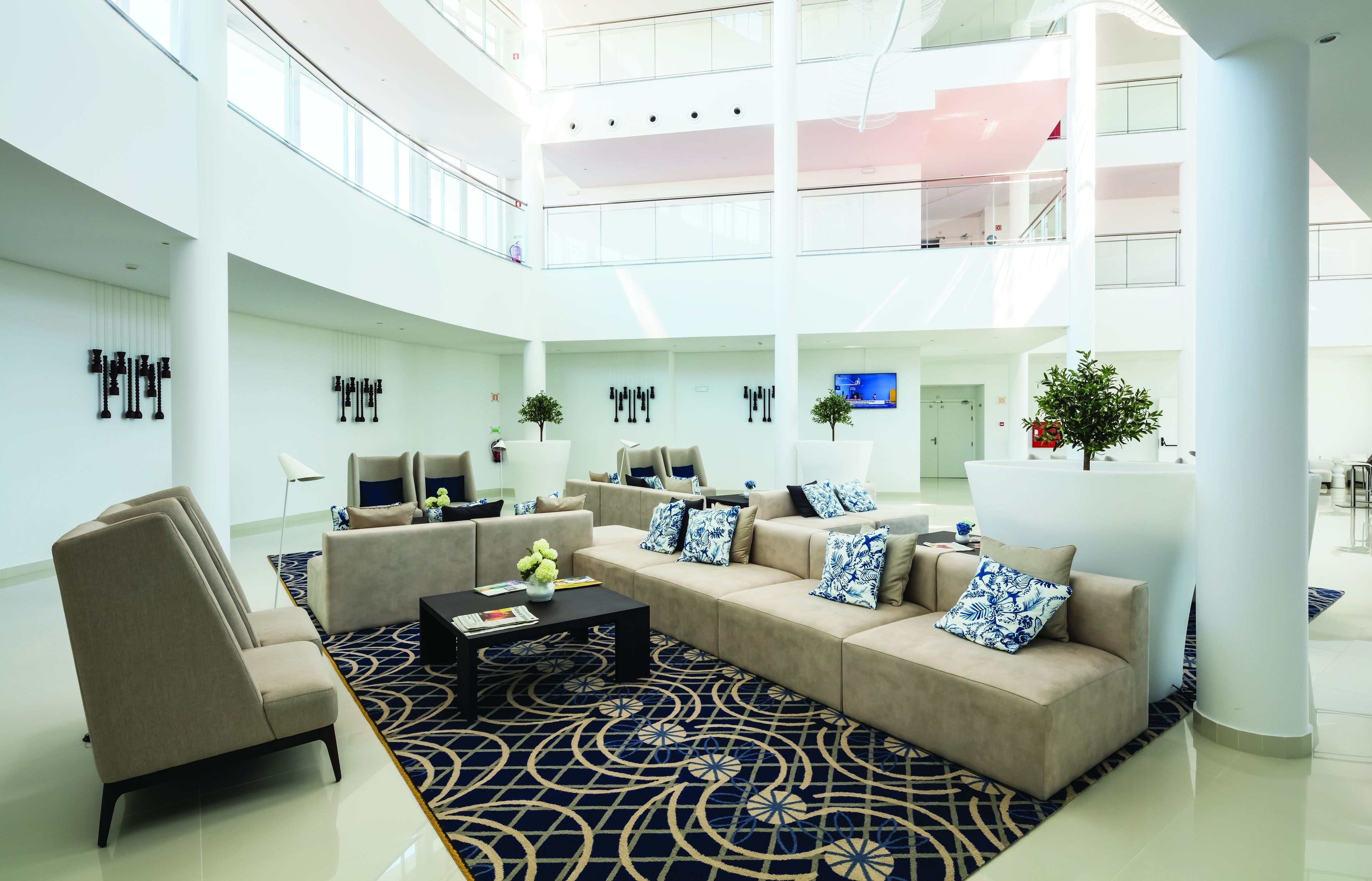 A spacious, modern lobby with a high ceiling and multiple levels. It features a large seating area with sofas and armchairs, a coffee table, and decorative plants.
