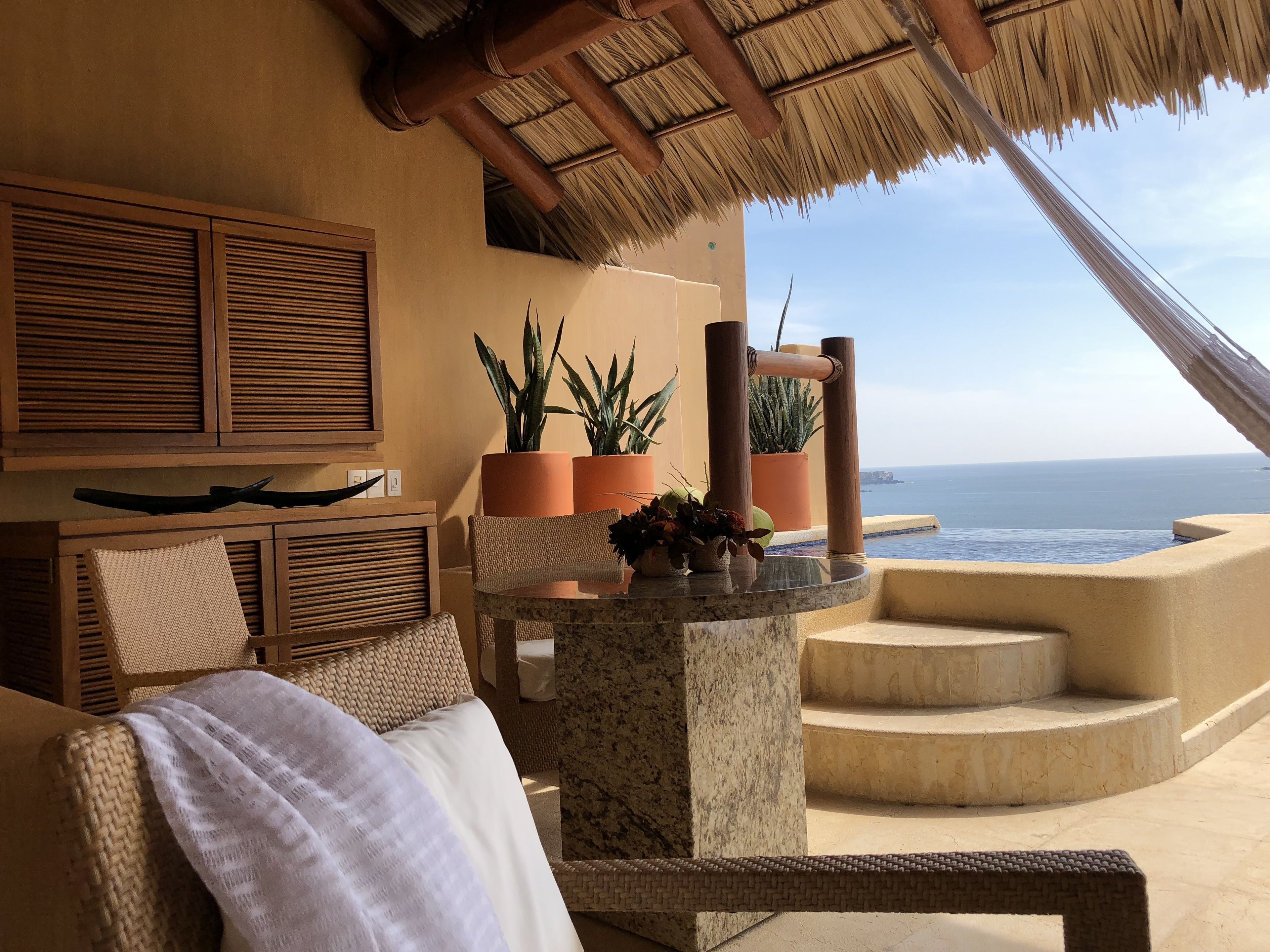 A balcony with a view of the ocean, featuring a thatched roof, wicker furniture, and potted plants.