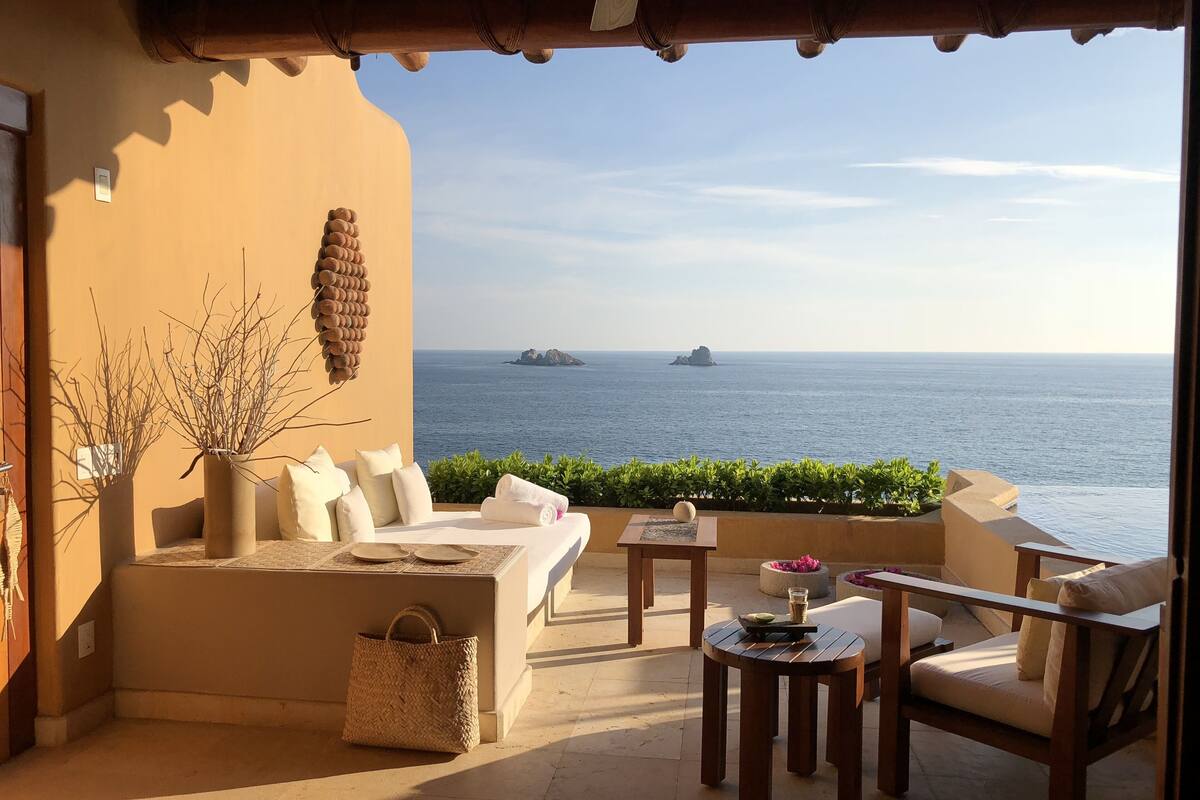 Een zonnig terras met uitzicht op zee, met een bank, stoelen, een tafel en een decoratief wandobject.