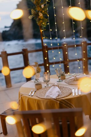 A table set for dining with a view of the sea, decorated with string lights and a vase of flowers.