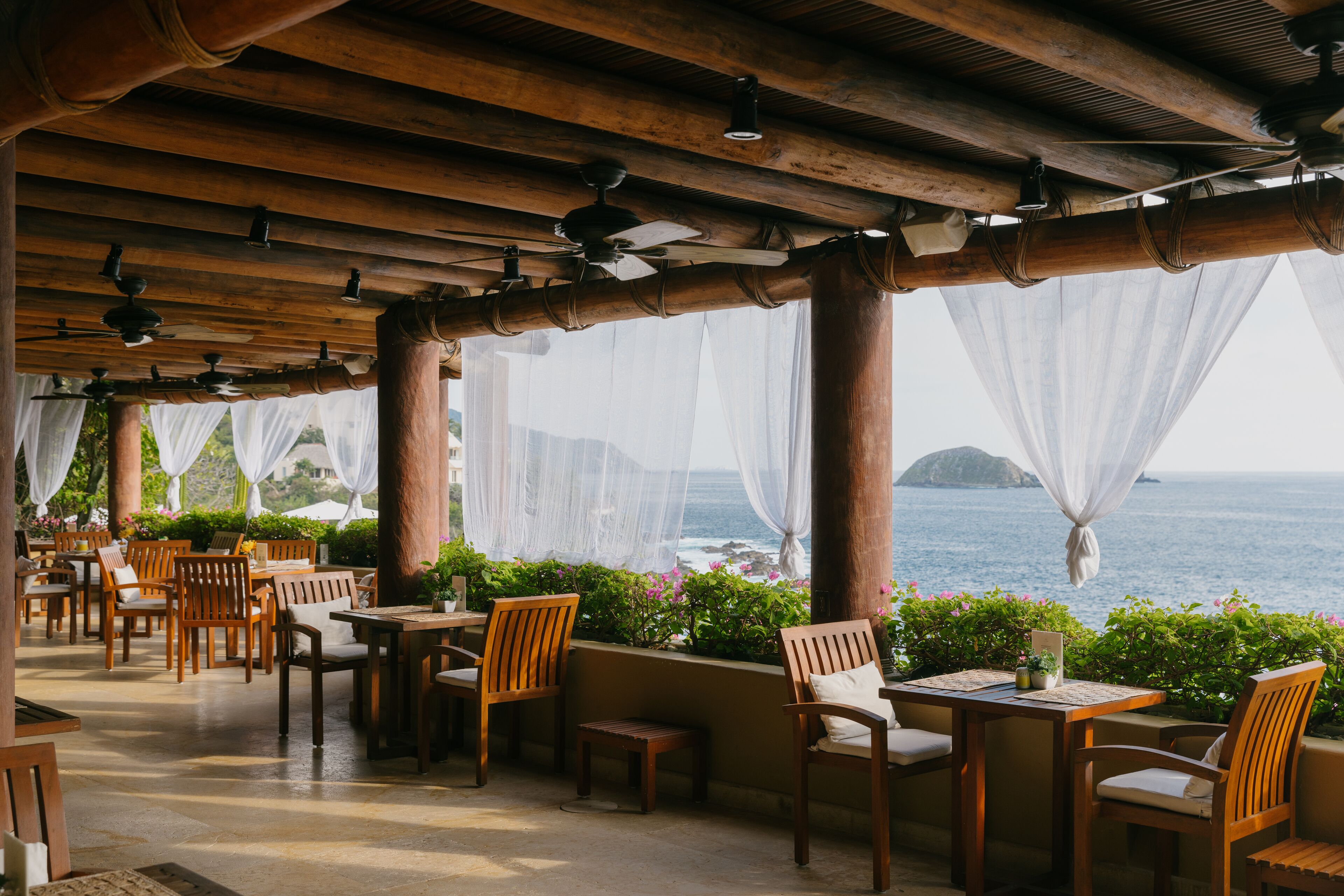 An outdoor dining area with wooden tables and chairs, covered by a wooden roof with ceiling fans, overlooking a body of water.