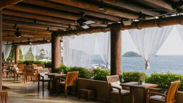 An outdoor dining area with wooden tables and chairs, covered by a wooden roof with ceiling fans, overlooking a body of water.