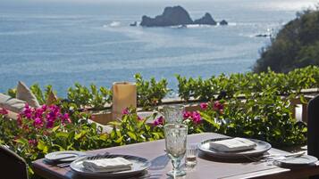 Mesa lista con platos, cubiertos y una copa, con vistas a la costa, flores y una vela.
