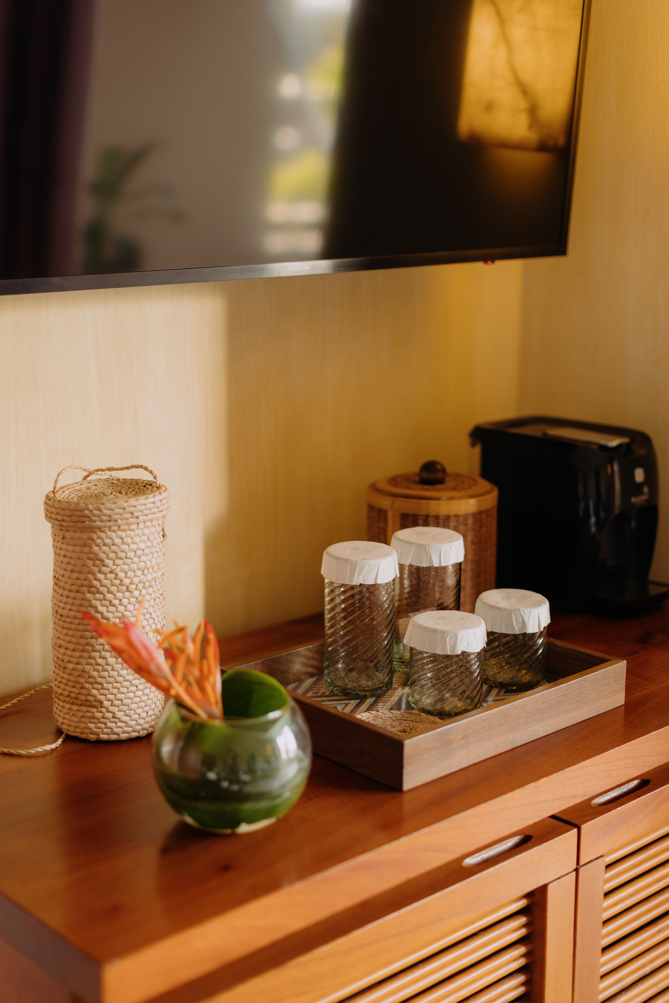 Una consola de madera con una cesta tejida, un florero de vidrio con flores secas y tres frascos con tapas. Un televisor está montado en la pared de arriba.