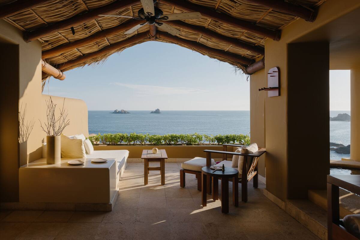 Een terras met rieten dak, een bank, een tafel en stoelen, met uitzicht op de zee en verre schepen.
