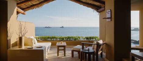 A terrace with a thatched roof, a sofa, a table, and chairs, overlooking the sea with distant ships.