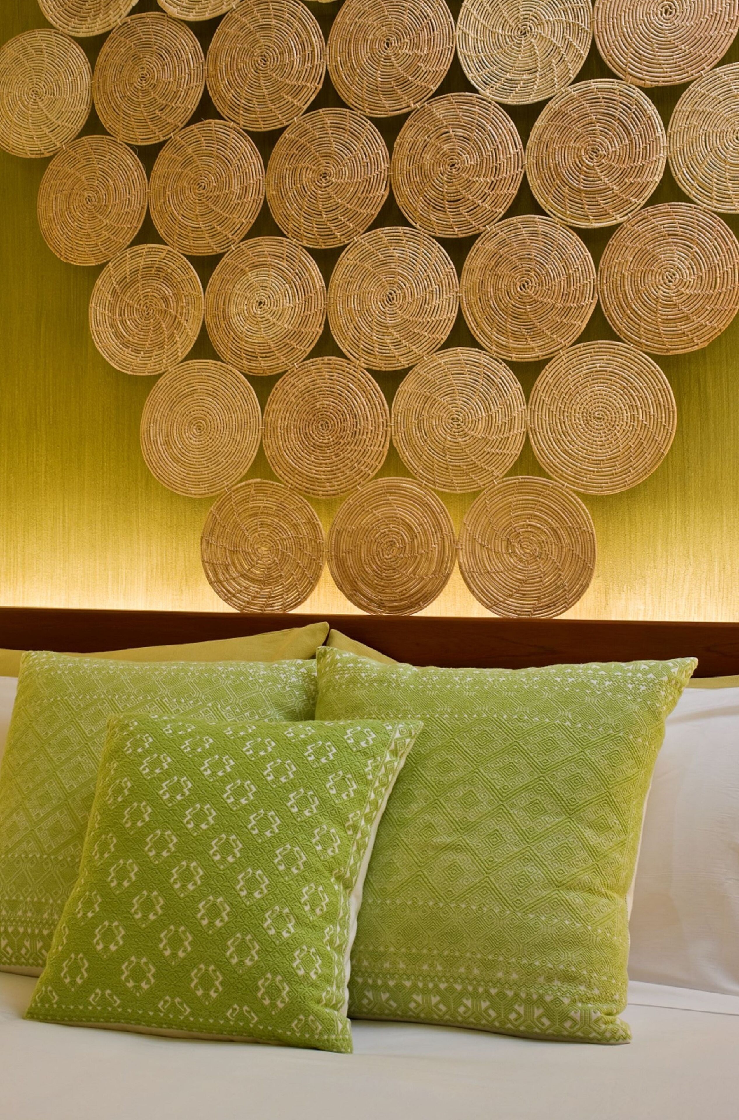 Una habitación con un panel circular tejido en la pared y almohadones estampados de color verde sobre la cama.