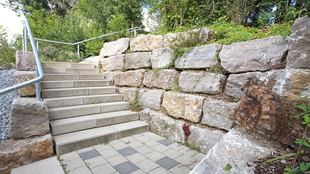 Unos escalones de piedra conducen a una terraza de madera, rodeada de vegetación.