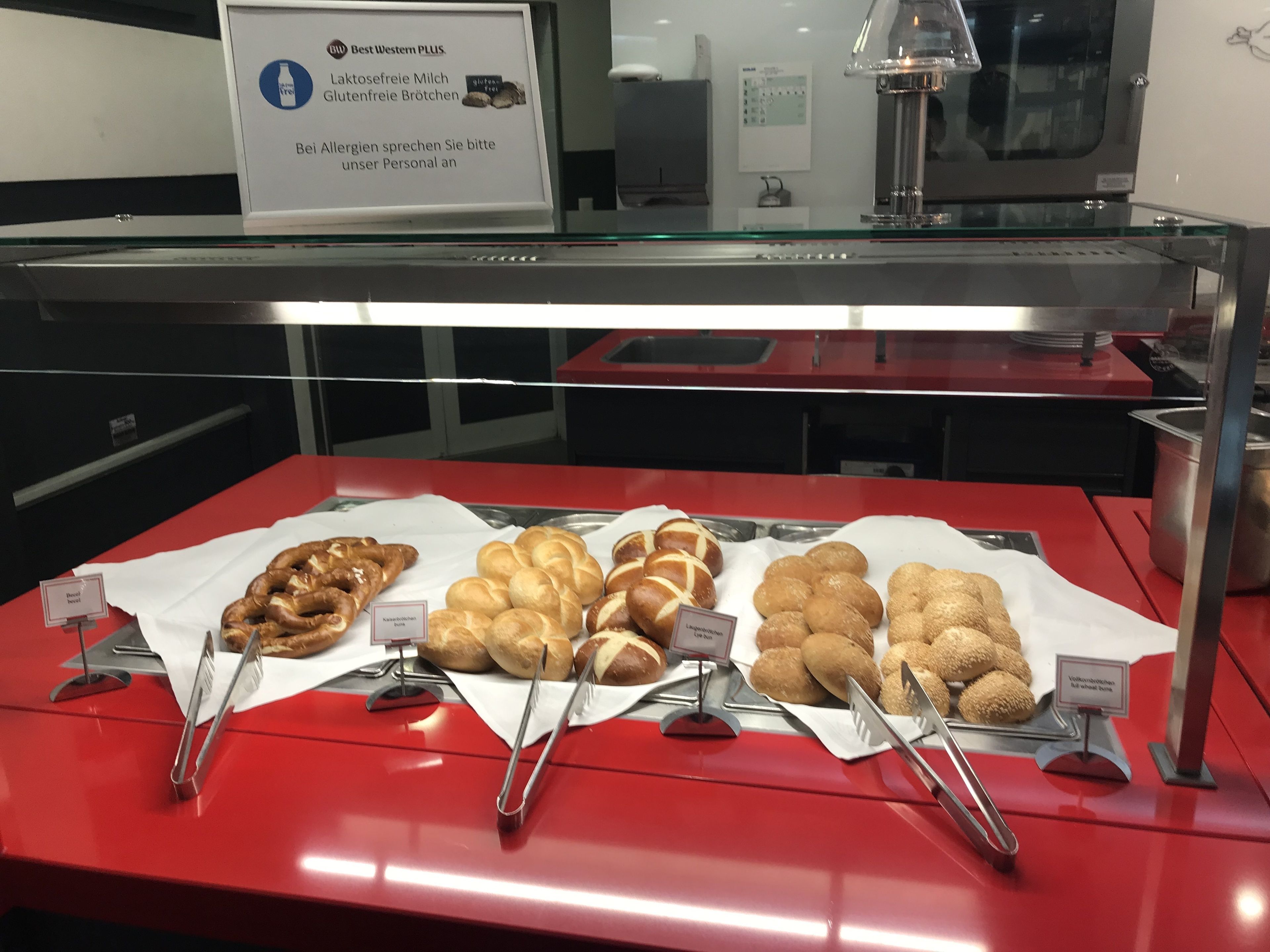 Mostrador de panadería con diferentes panes y pasteles, incluyendo pretzels, croissants y bollos, con etiquetas que indican sus nombres.