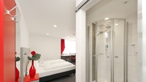 Una habitación de hotel moderna con ducha de vidrio, una cama con ropa blanca y un florero rojo con una flor.