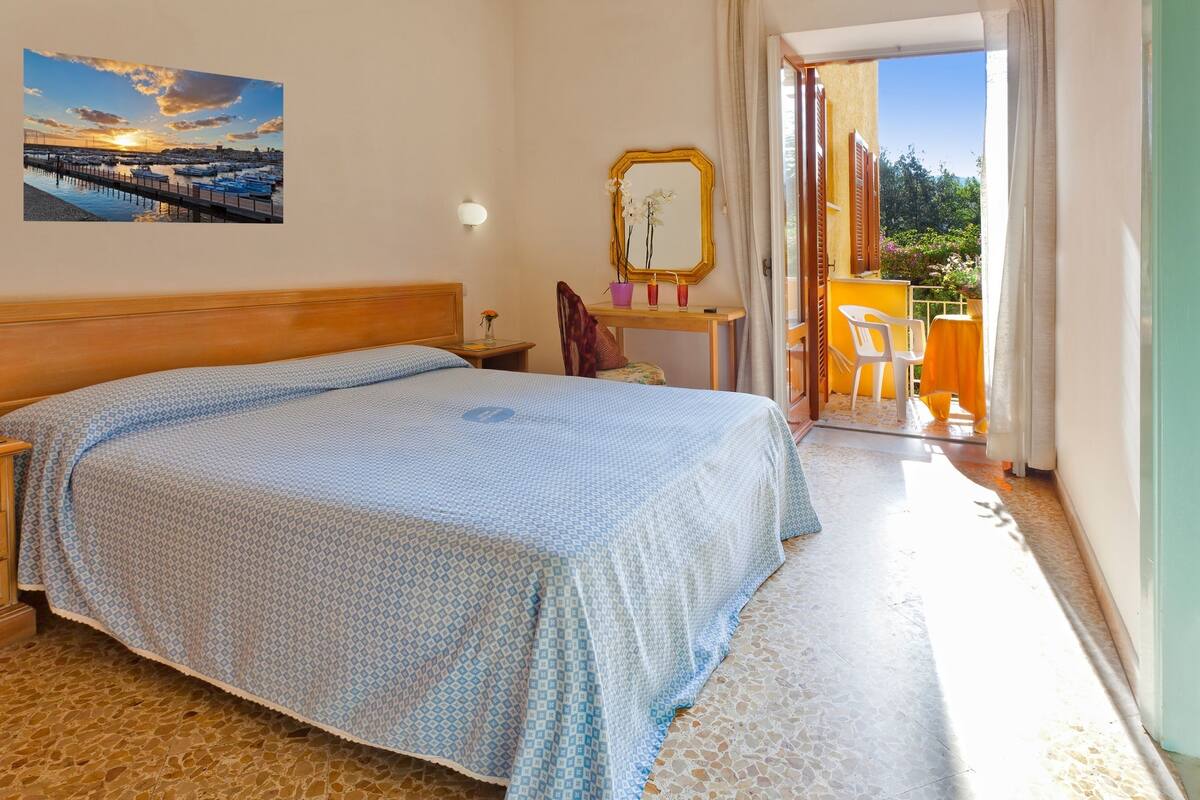 Un dormitorio con cama, mesita de noche, una pequeña mesa con un florero, espejo y vistas a un balcón con sillas.