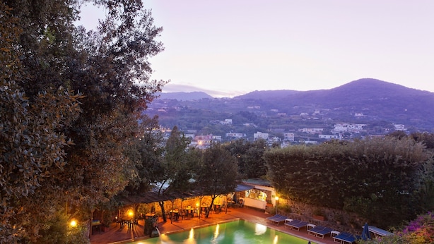 A swimming pool with lounge chairs and a covered seating area, surrounded by trees and overlooking a town. - Hotel Cleopatra (Ischia)