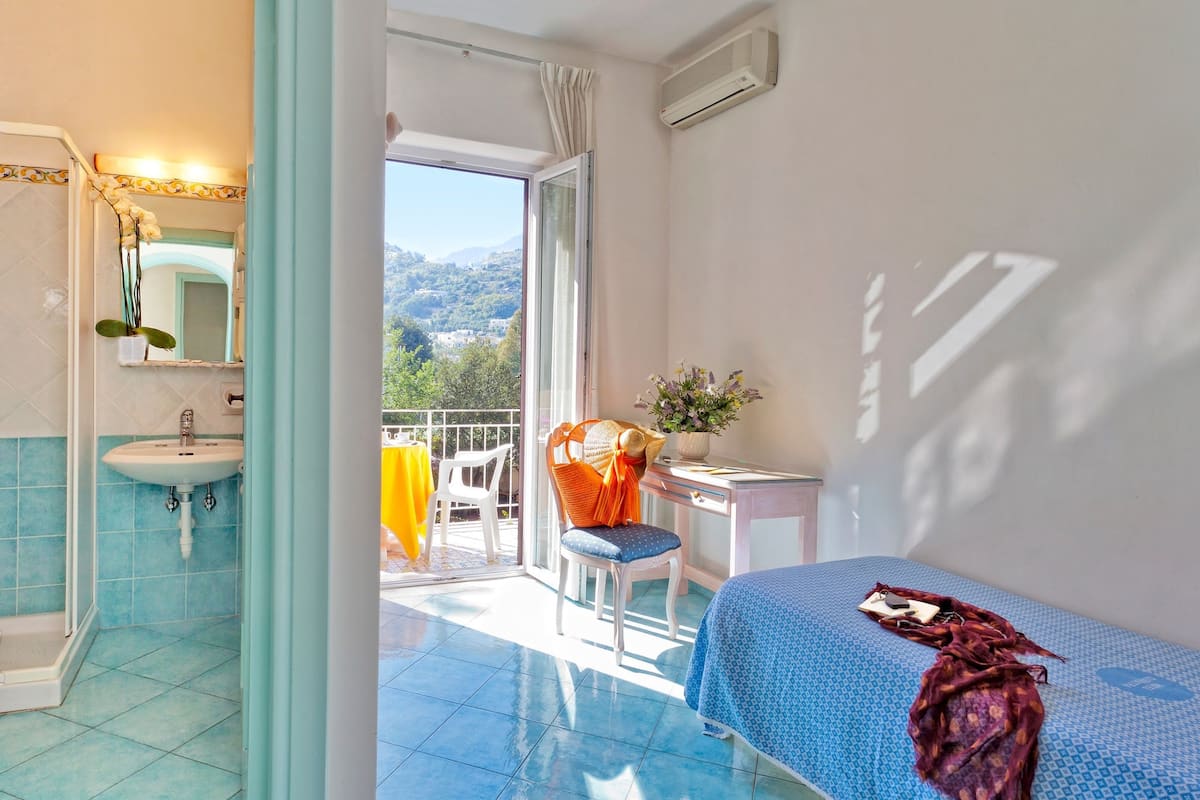 Una habitación con cama, escritorio, silla, balcón con vistas al campo y baño con lavabo y espejo.
