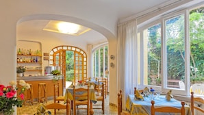 A restaurant with wooden tables and chairs, a bar area with shelves of bottles, and large windows offering a view of greenery. - Hotel Cleopatra (Ischia)