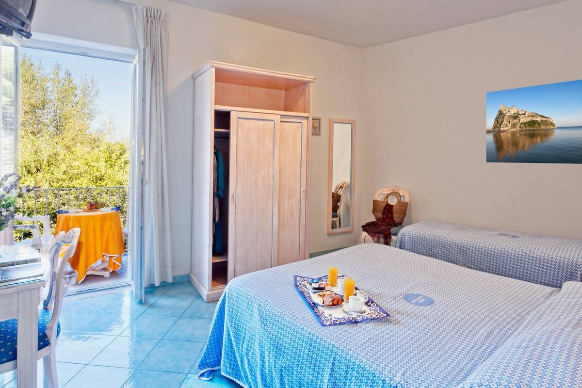 Habitación de hotel con dos camas, un balcón con mesa y sillas, un armario y un cuadro de un paisaje costero.