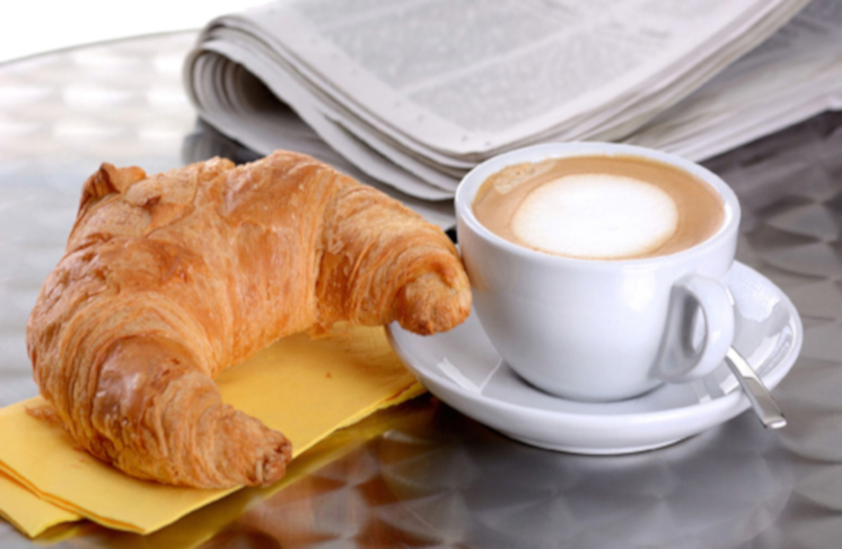 A croissant and a cup of coffee on a glass table with newspapers.