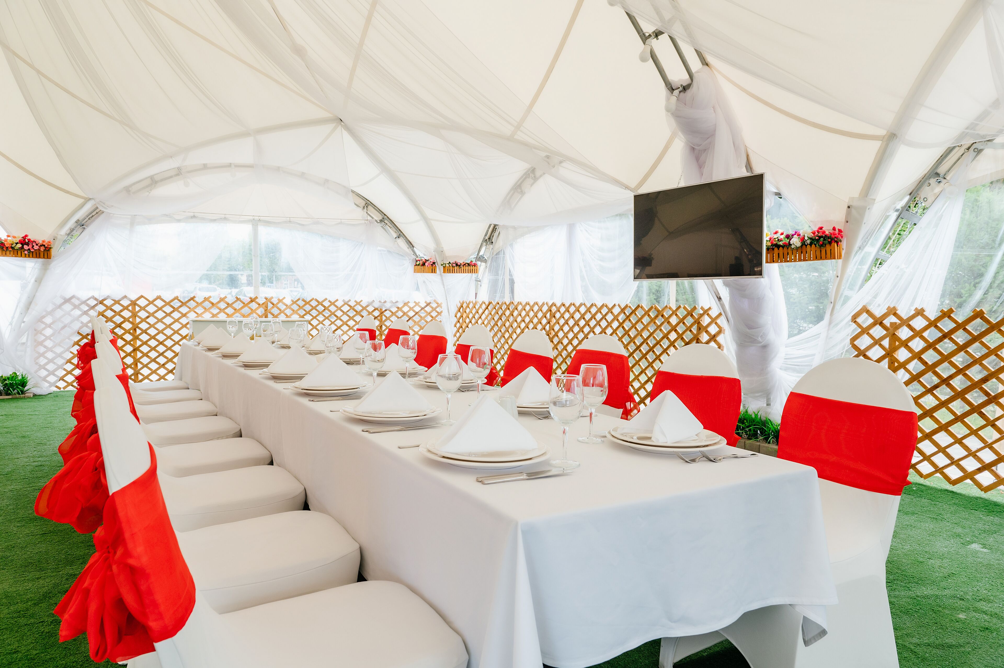 A tented dining area with white tables, red sashes, and a television screen.