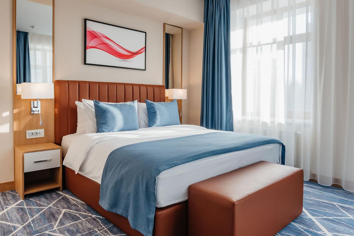 Une chambre d’hôtel comprenant un lit, une table de chevet, une lampe, un miroir, une fenêtre avec des rideaux et une œuvre d’art accrochée au mur.